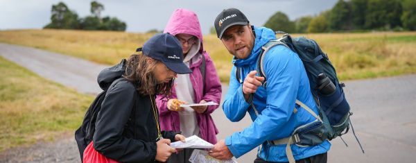 An Out There Award instructor wearing a Ramblers cap points ahead while two young people stand beside him on a path, holding maps and checking their route in open countryside.